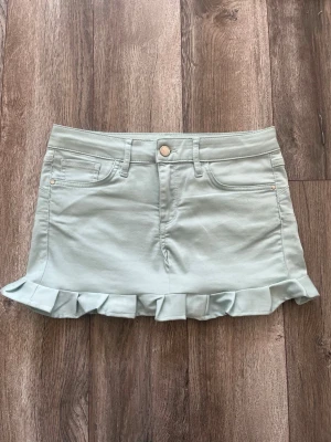 Ljusgrön volangkjol med fickor - Supersöt ljusgrön kjol med volangkant längst ner och klassiska jeansfickor både fram och bak. Kjolen har bälteshällor och stängs med knapp och dragkedja. Perfekt för dig som gillar en lekfull och trendig stil.