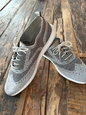 Grå stickade sneakers från Cole Haan - Snygga grå sneakers från Cole Haan med stickad ovandel och vita sulor. Skorna har klassisk snörning och en låg profil. Perfekta för dig som gillar en stilren och modern look med bekvämt material och snygga detaljer. Storlek 40,5