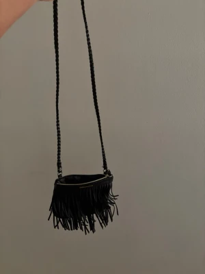 Svart axelväskor - Säljer en svart axelväska från Saddler med långa fransar framtill och flätad axelrem. Väskan har guldfärgad dragkedja och logga på framsidan. Perfekt för dig som gillar boho-stil och vill ha en unik accessoar till din outfit. Säljer också en till väska tillsammans med denna, en basic svart som är så fin och passar till allt 