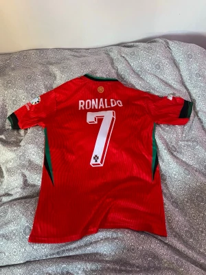 Portugal Ronaldo matchtröja Nike - Säljer en röd Portugal fotbollströja från Nike med nummer 7 och Ronaldo på ryggen. Tröjan har gröna detaljer vid krage och ärmslut, samt officiella tryck och loggor. Tillverkad i lätt och ventilerande Dri-FIT material, perfekt för träning eller match.