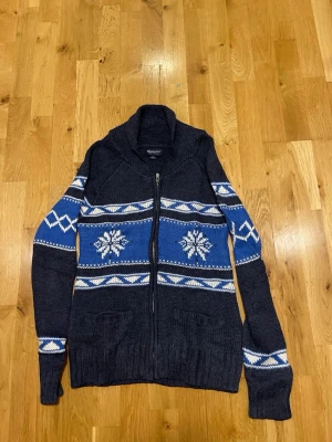 Mörkblå stickad kofta med dragkedja stl L - Mysig mörkblå stickad kofta från Bluenotes med dragkedja framtill. Har blå och vita mönster med snöflingor och geometriska detaljer över bröstet och ärmarna. Två fickor framtill och ribbstickade muddar. Perfekt för kalla dagar.