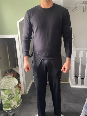 Svart sweatshirt från C.P. Company - Svart sweatshirt från C.P. Company med rund halsringning och ribbade muddar. Tröjan har en ikonisk linsdetalj på vänster ärm och är tillverkad i mjukt bomullsmaterial. Perfekt för en avslappnad och stilren look.