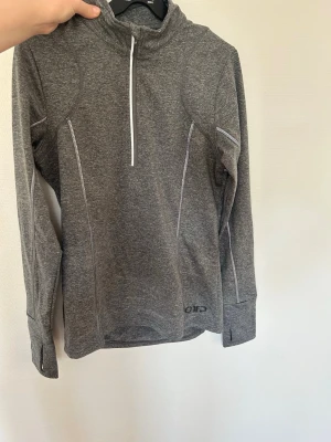 Grå träningströja med halv dragkedja OTP - Sportig grå långärmad träningströja  med halv dragkedja framtill och vita kontrastsömmar. Tillverkad i ett stretchigt funktionsmaterial som passar perfekt för löpning eller gym. Tumhål vid ärmsluten för extra komfort.