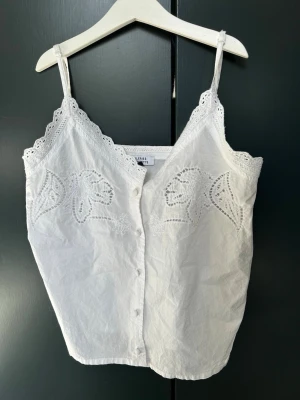Vit broderad linnetopp med spets - Söt vit linnetopp från Lindex med smala axelband, spetsdetaljer längs kanterna och broderade blommor framtill. Toppen har knäppning framtill och är tillverkad i bomull, perfekt för varma dagar.