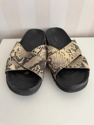 Slip-in sandaler med ormskinnsmönster - Snygga slip-in sandaler från Rizzo med breda korslagda remmar i beige och svart ormskinnsmönster. Sulan är svart och har en lätt platå. Perfekta för en trendig och avslappnad look. Remmarna är i syntetmaterial och sulan är i gummi.Storlek 37-38