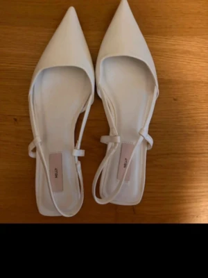 Vita ballerinaskor med spetsig tå - Snygga vita ballerinaskor från Nelly med spetsig tå och smala remmar runt hälen. Skorna har en stilren design och är tillverkade i ett slätt material som ger en modern look. Perfekta för dig som gillar minimalistisk stil.