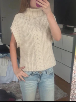 Beige stickad ärmlös polotröja - Säljer en beige stickad ärmlös polotröja med grovt kabelstickat mönster framtill.