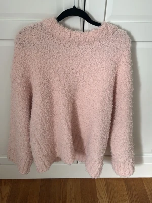 Ljusrosa fluffig stickad tröja - Supermjuk och fluffig stickad tröja i ljusrosa färg. Tröjan har en avslappnad passform med rund hals och långa ärmar. Perfekt för att hålla sig varm och mysig under kyliga dagar.