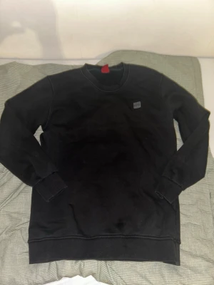 Svart sweatshirt från Hugo boss - En stilren svart sweatshirt från hugo boss med rund halsringning och diskret logopatch på bröstet. Tröjan har långa ärmar och ribbade muddar vid ärmslut och nederkant. Perfekt för en avslappnad och enkel look.