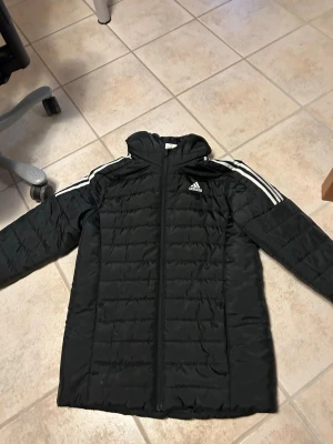 Svart pufferjacka från Adidas - Svart pufferjacka från Adidas med huva och dragkedja framtill. Klassiska vita ränder längs axlarna och Adidas-logga på bröstet. Jackan är lång och har en sportig look, perfekt för kalla dagar. Tillverkad i vadderat material för extra värme.
