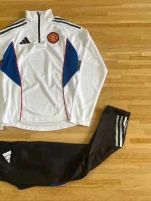 Manchester United Adidas träningsset - Snyggt träningsset från Adidas med Manchester United-emblem. Överdel i vitt med blå och röda detaljer, halv dragkedja och svarta Adidas-ränder på axlarna. Matchande svarta byxor med vita ränder och Adidas-logga. Perfekt för fotbollsträning eller gym.