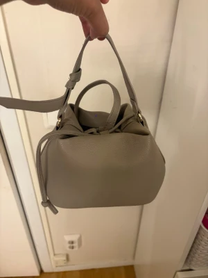 Beige bucket bag med axelrem - Stilren beige bucket bag i mjukt skinnliknande material med både handtag och justerbar axelrem. 