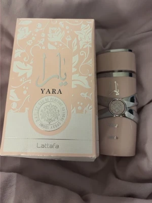 Yara parfym från Lattafa, rosa - Yara parfym från Lattafa i en snygg rosa flaska med silverdetaljer och matchande ask. Flaskan har en unik trekantig form och dekorativa mönster. Doften är Eau de Parfum och designen känns både modern och elegant, perfekt för dig som gillar stilrena accessoarer. Doftar vanilj, söt, pudrig och lite blommig.