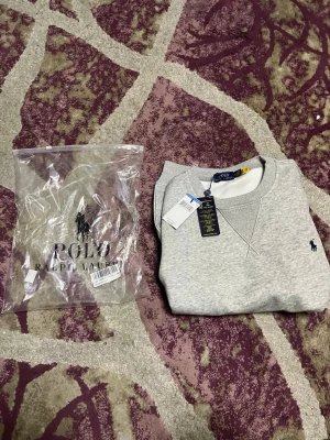 Grå stickad tröja från Polo Ralph Lauren - Snygg grå stickad tröja från Polo Ralph Lauren med klassisk rund halsringning och ribbade muddar. Tröjan har den ikoniska mörkblå logotypen broderad på bröstet och är tillverkad i mjukt material för en clean och stilren look.