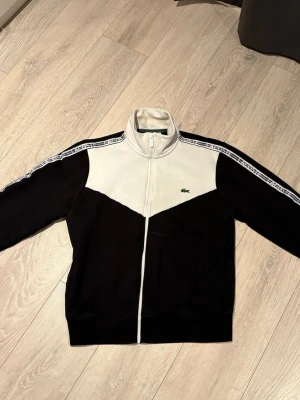 Svartvit zip hoodie från Lacoste - Snygg svartvit hoodie från Lacoste med dragkedja framtill och hög krage. Ikoniska Lacoste-loggan på bröstet och vita band med Lacoste-tryck längs axlarna. Tillverkad i mjukt material, perfekt för en chill och sportig look.