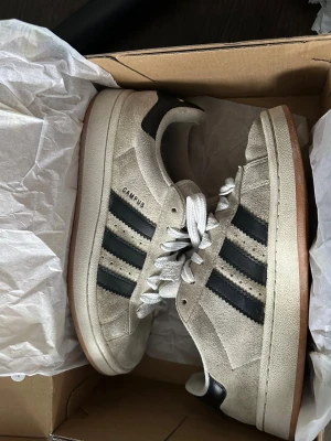 Adidas Campus beige sneakers - Säljer ett par Adidas Campus sneakers i beige mocka med svarta klassiska stripes på sidorna och vit sula. Skorna har vita skosnören och gummisula i brunt. Perfekta för dig som gillar retrostil och streetwear.