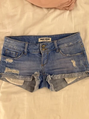 Blå jeansshorts från Tally Weijl - Säljer ett par blå jeansshorts från Tally Weijl med slitna detaljer och upprullade benslut. Shortsen har fem fickor, dubbla knappar och klassisk jeanslook. Perfekta för varma dagar och en avslappnad stil.