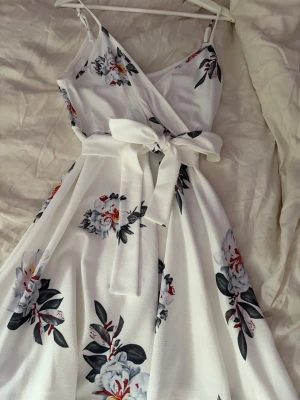 Vit blommig omlottklänning med knyt - Säljer en vit omlottklänning med blommigt mönster i blått, rött och grått. Klänningen har smala axelband, v-ringning och ett brett midjeband  som går att ta bort och som knyts i en rosett. Materialet känns lätt och luftigt, perfekt för varma dagar.