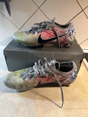 Nike Mercurial vapor 13 elite neymar jr  - Säljer ett par Nike Mercurial fotbollsskor med grå bas och färgstänk i gult, rött och blått. Skorna har svart Nike-swoosh på sidan, snörning och dobbar för spel på gräs. Ovandelen är i syntetmaterial och designen är riktigt iögonfallande för dig som vill sticka ut på planen.