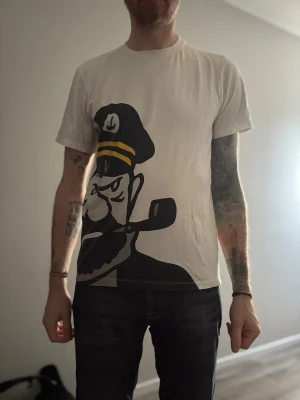 Vit t-shirt med stor tryckt kaptendesign - Säljer en vit t-shirt med en stor, grafisk kaptendesign i svart och gult framtill. T-shirten har korta ärmar och normal passform. Perfekt för dig som gillar unika och uttrycksfulla plagg.