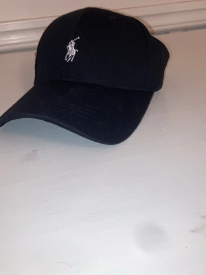 Svart keps från Polo Ralph Lauren - Snygg svart keps från Polo Ralph Lauren med den klassiska broderade loggan i vitt framtill. Justerbar baktill för perfekt passform och tillverkad i mjukt bomullstyg. Enkel och stilren design som passar till det mesta.