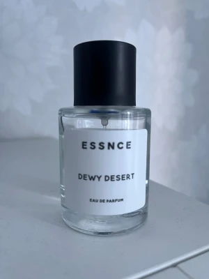 ESSNCE Dewy Desert Eau de Parfum - Minimalistisk parfymflaska från ESSNCE med doften Dewy Desert. Flaskan är cylindrisk i genomskinligt glas med en enkel vit etikett och svart rund kork. Modern och stilren design som passar perfekt på parfymhyllan.
