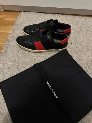 Svarta Saint Laurent sneakers med röd detalj - Skicka erbjudande. Snygga svarta sneakers från Saint Laurent med röda detaljer på sidan och hälen. Skorna har en klassisk låg profil, svarta skosnören och vit sula. Tillverkade i slätt läder för en stilren look. Perfekta för dig som gillar exklusiva märken och enkel design.
