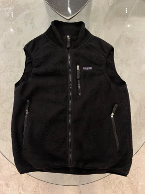 Svart fleeceväst från Patagonia - Säljer en svart fleeceväst från Patagonia med dragkedja framtill och två sidofickor med dragkedja. Västen har en liten bröstficka med dragkedja och klassisk Patagonia-logga på bröstet. Tillverkad i mjuk fleece som håller dig varm och bekväm.