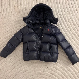 Svart dunjacka Polo Ralph Lauren - Snygg svart dunjacka från Polo Ralph Lauren, storlek 16 y/xl (barn) med huva och dragkedja framtill. Jackan har två fickor med dragkedja och den klassiska röda loggan broderad på bröstet. Perfekt för kalla vinterdagar och riktigt stilren look.
