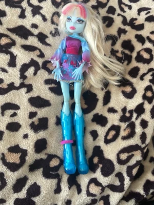Monster High docka Abbey - Säljer en Monster High docka med långt blont hår, blå hud och stora blå stövlar. Dockan har en färgglad outfit med rosa topp och blå kjol med mönster. Perfekt för samlare eller lek.