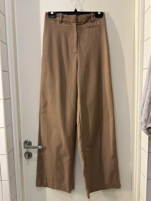 Beiga vida kostymbyxor - Säljer ett par beiga kostymbyxor med hög midja och vida ben. Byxorna har bälteshällor, dold dragkedja och en dekorficka fram. Materialet känns mjukt och har en lätt struktur, perfekt för en clean och stilren look.