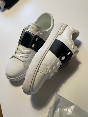 Valentino vita sneakers med svart detalj - Snygga vita sneakers från Valentino med bred svart läderdetalj över sidan och silverfärgade nitar. Klassisk låg modell med vita skosnören och robust sula. Perfekta för dig som vill ha en stilren och trendig look.
