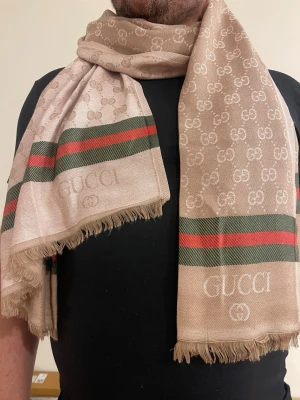 Beige halsduk från GG med logga - Snygg beige halsduk från  med klassiskt GG-mönster och gröna samt röda ränder. Halsduken har fransade kanter och -loggan invävd på båda sidor. Perfekt accessoar för att lyfta din outfit med en lyxig touch.