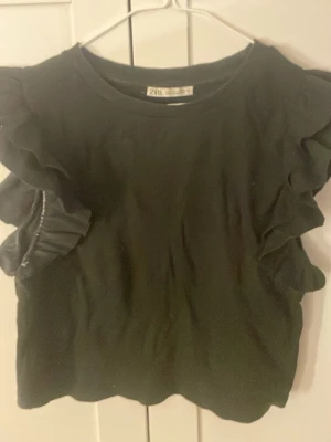 Svart volangtopp från Zara - Snygg svart topp från Zara med volangdetaljer längs ärmarna. Toppen har rund halsringning och är tillverkad i ett mjukt material som känns bekvämt mot huden. Perfekt för dig som vill ha en enkel men ändå trendig look.