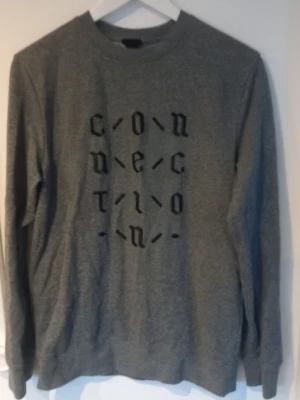 Grå sweatshirt med tryck - Grå långärmad sweatshirt med svart grafiskt tryck på bröstet där det står 'connection' i ett coolt typsnitt. Tröjan har rund halsringning och ribbade muddar vid ärmslut och nederkant. Perfekt för en avslappnad streetstyle-look.