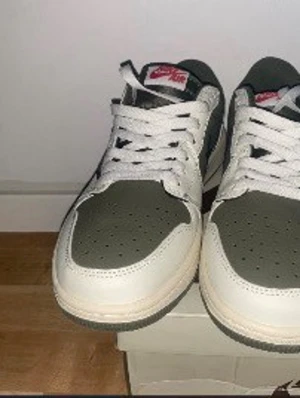 Nike Air Jordan 1 Low Travis Scott - Nike Air Jordan 1 Low Travis Scott i vitt och olivgrönt läder med ikonisk omvänd Swoosh och röda detaljer på tungan. Skorna har perforerad tåbox och kommer med extra skosnören i beige och oliv. Perfekt för dig som gillar streetwear och unika sneakers.