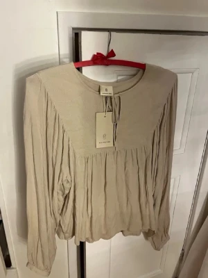 Beige blus från Peppercorn - Luftig beige blus från Peppercorn med rund halsringning och dekorativ knytning framtill. Tillverkad i ett mjukt, strukturerat tyg som ger en avslappnad och trendig look. Perfekt att styla med jeans eller kjol för en enkel men snygg outfit.