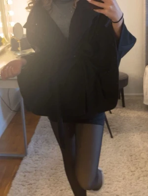 Svart kappa med knytband - Stilren svart kappa med brett knytband i midjan och vida ärmar. Kappan har en oversized passform och går ner till låret. Perfekt för dig som vill ha en trendig och enkel look. Passar till lager på lager och funkar till många outfits.
