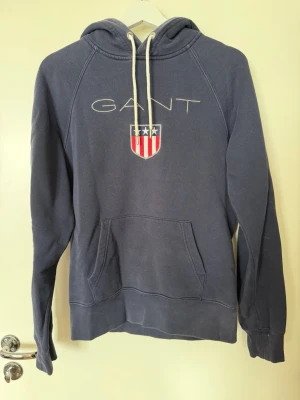 Mörkblå hoodie från GANT med logga - Snygg mörkblå hoodie från GANT med vit broderad logga och sköld på bröstet. Klassisk känguruficka framtill och vita dragsnören i huvan. Tillverkad i mjuk bomullsblandning, perfekt för en avslappnad och sportig stil.