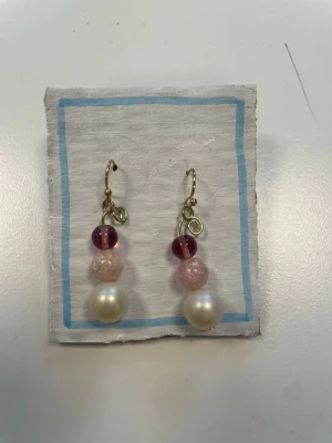 Hängande örhängen med pärlor och rosa detaljer - Säljer ett par unika hängande örhängen med guldfärgade krokar. Örhängena har tre pärlor vardera: en stor vit pärla längst ner, en ljusrosa pärla i mitten och en mörkrosa/glasklar pärla högst upp. Perfekt för dig som gillar färg och statement-accessoarer.