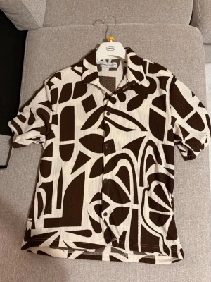 Brunvit mönstrad skjorta från Bershka - Kortärmad skjorta från Bershka i brun och vit med ett grafiskt, rutigt mönster. Skjortan har klassisk krage, knappar framtill och är gjord i ett strukturerat bomullstyg. Perfekt för dig som vill sticka ut med en unik look.