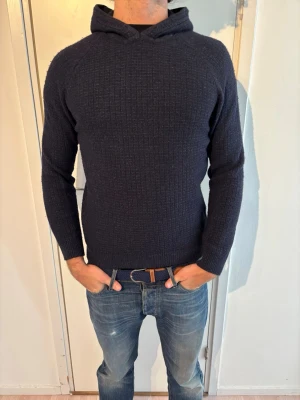 Massimo Dutti hoodie - Säljer en mörkblå stickad hoodie från Massimo Dutti med lång ärm och ribbade muddar. Tröjan har en diskret struktur i stickningen och en klassisk huva. Perfekt för dig som gillar en enkel men snygg stil. Materialet känns mjukt och bekvämt.