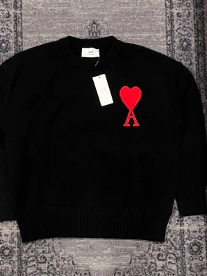 Svart stickad tröja från Ami - Svart stickad tröja från Ami Alexandre Mattiussi med röd hjärtlogga och bokstaven A på bröstet. Tröjan har rund halsringning, ribbade muddar och är gjord i mjukt material. Perfekt för dig som gillar stilrena detaljer och vill ha något extra i garderoben.