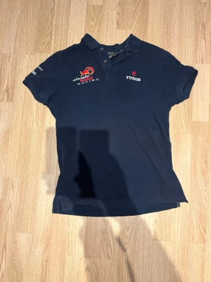 Marinblå polotopp sail Racing - Snygg marinblå polotopp från sail Racing med broderade loggor och text på bröstet och ärmen. Klassisk pikékrage med knappar framtill och korta ärmar. Perfekt för dig som gillar motorsport och vill ha en sportig look.