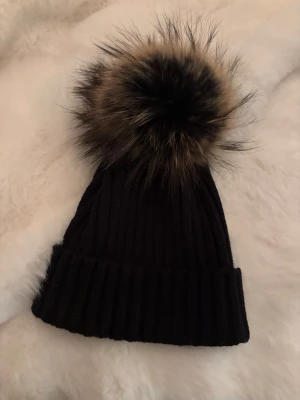 Svart ribbstickad mössa med pälsboll - Snygg svart ribbstickad mössa med stor fluffig pälsboll i bruna och svarta toner. Mössan har uppvikt kant och är perfekt för kalla dagar. Materialet ser ut att vara mjukt och stretchigt, vilket gör den bekväm att bära.