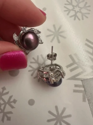 Silvriga örhängen med mörka pärlor - Säljer ett par eleganta örhängen i silverfärgad metall med mörklila pärlor och små gnistrande stenar. Designen är blommig och detaljerad, perfekt för dig som gillar unika accessoarer. Stiftmodell som sitter säkert på örat.