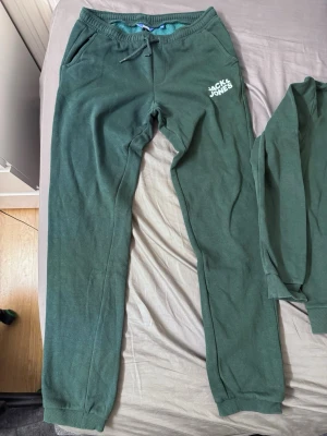 Grönt loungewear-set Jack & Jones - Mysigt loungewear-set från Jack & Jones i mörkgrönt. Setet består av mjuka byxor med dragsko och fickor samt en matchande hoodie med tryck framtill. Perfekt för chill dagar hemma. Materialet känns bomullsliknande och plaggen har avslappnad passform.