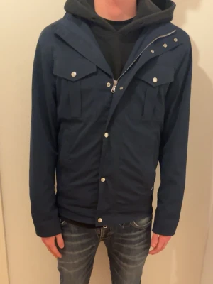 Mörkblå vindjacka med tryckknappar - Snygg mörkblå vindjacka med tryckknappar och dragkedja framtill. Jackan har två bröstfickor med lock och lång ärm. Perfekt för dig som vill ha en stilren och enkel jacka till vardagslooken.skriv om undringar 