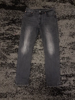 Levi’s 501 - Säljer ett par grå straight 501 jeans från Levi's med klassisk femficksdesign och diskret slitning på framsidan. Jeansen har normal midja och är tillverkade i denim. Perfekta för en avslappnad och trendig look.