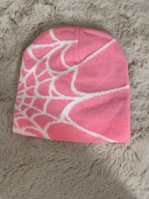 Rosa mössa med spindelnätsmönster - Säljer en rosa mössa med vitt spindelnätsmönster. Mössan är mjuk och stretchig, perfekt för dig som vill sticka ut lite extra. Materialet känns som bomull eller akryl och designen är riktigt cool och ungdomlig.